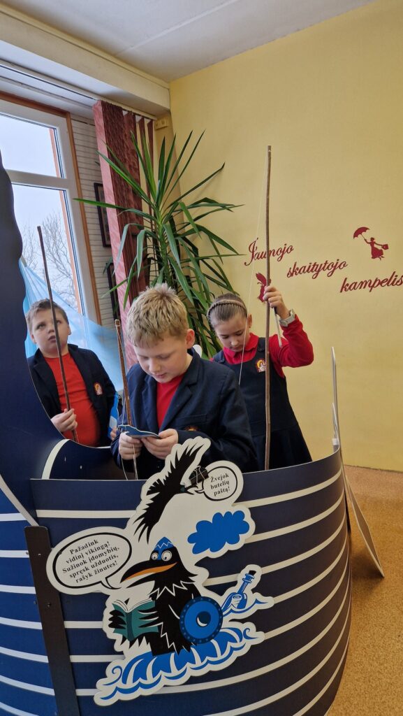 Trys mokiniai stovi dekoratyviame mėlyname „Vikingų laivo“ makete ir laiko meškeres, atlikdami edukacinę veiklą. Vienas mokinys skaito kortelę, kiti stebi savo „laimikį“. Ant sienos matyti užrašas „Jaunojo skaitytojo kampelis“, šalia auga didelis kambarinis augalas, pro langą krinta dienos šviesa.