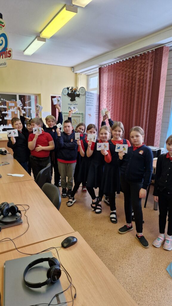 Grupė pradinių klasių mokinių stovi klasėje ir laiko pakeltas mažas korteles su piešiniais; priekyje matyti stalai su kompiuteriais ir ausinėmis, o fone – informacinis stendas ir langas su užuolaidomis.