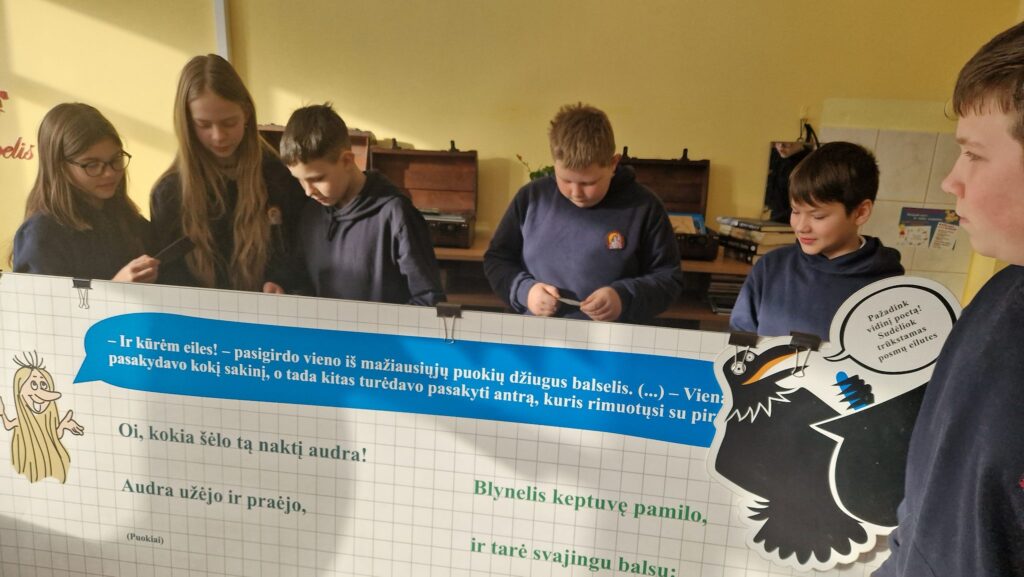 Penki mokiniai klasėje stovi prie edukacinio stendo su eilėraščio tekstu ir iliustracija, rankose laiko korteles bei atlieka užduotį; fone matyti knygos ir patefonai ant lentynos.