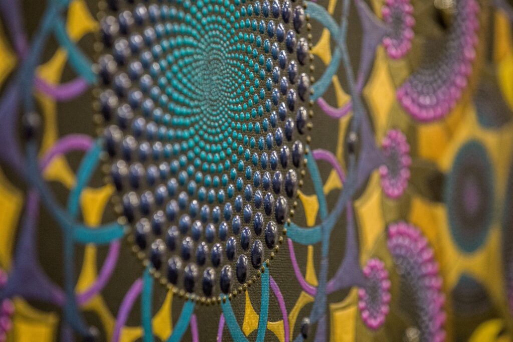 Paveikslėlyje matome artimą detalių nuotrauką, kurioje vaizduojama geometrinė struktūra su ryškiomis spalvomis. Mandala sudaryta iš taškų, kurie sukuria spiralinius raštus ir griežtai išdėstytus geometrinius motyvus. Dominuoja mėlyna, žalia, violetinė ir geltona spalvos, kurios harmoningai dera tarpusavyje. Paveikslas atrodo dinamiškai ir spalvingai, su akcentu į smulkias detales, kurios suteikia kūriniui gilumo ir tekstūros.