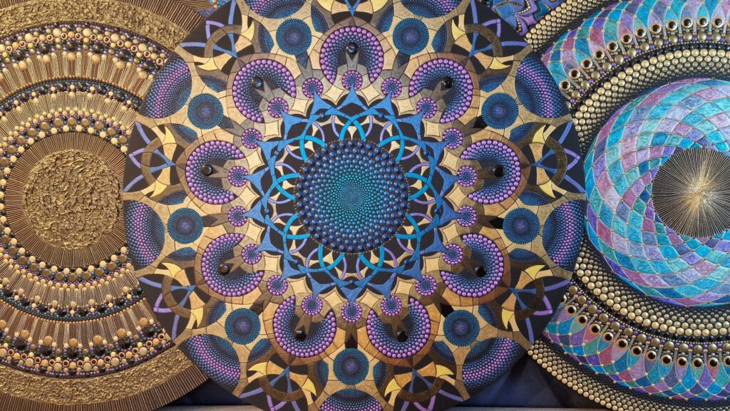 Paveikslėlyje matome trijų mandalų tipo paveikslų detalę, kurių kiekvienas turi unikalų, labai detaliai išpieštą geometrinį raštą. Pirmas paveikslas, esantis kairėje, turi subtilius auksinius ir mėlynos spalvos tonus, su ryškiais taškais ir tekstūromis. Antras paveikslas centre yra sudarytas iš įvairių geometrinių figūrų, apjungiančių mėlynas, violetines, ir auksines spalvas, o trečias paveikslas dešinėje – su išraiškingais, spiralės formos elementais, kuriuose atsispindi mėlyna, rožinė ir auksinė spalvos. Visi trys paveikslai harmoningai derinasi ir sukuria įspūdingą, dinamišką vizualinį efektą.