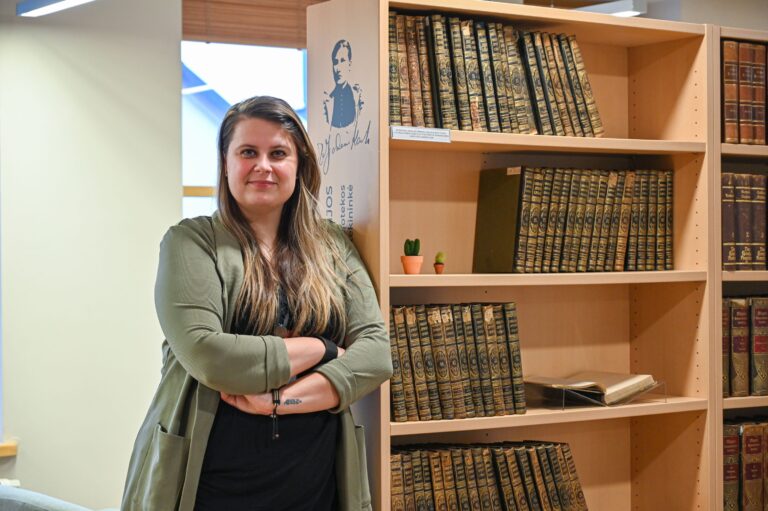 Greta Kėvelaitienė stovi sunėrusi rankas prie knygų lentynos bibliotekos skaityklos fone