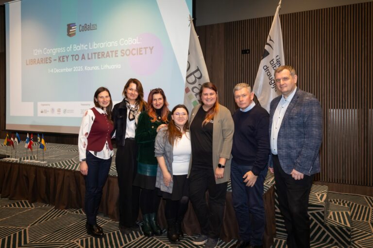 Septynių asmenų grupė renginio dalyvių projektoriaus fone salės aplinkoje