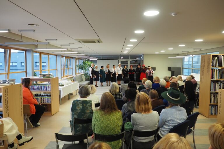 Grupė dalyvių sėdi priešais dainuojančią atlikėjų grupę bibliotekos skaityklos fone