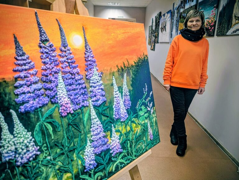 Moteris stovi galerijos koridoriuje šalia molberto pastatyto paveikslo, kuriame pavaizduotos violetinės lubinų gėlės saulėlydžio fone; ant sienų už jos kabo daugiau spalvingų paveikslų.