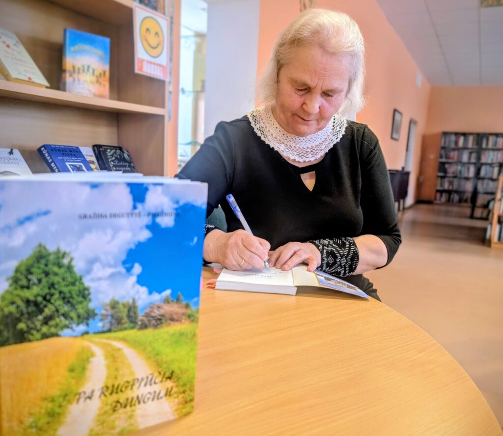 Bibliotekoje prie stalo sėdinti moteris juodais drabužiais su šviesia apykakle pasirašo knygą. Pirmame plane – knygos viršelis su lauko keliuko ir medžio vaizdu, fone – knygų lentynos.