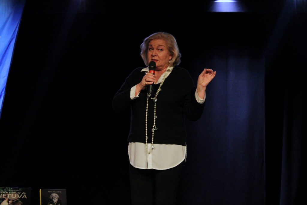 Edita Mildažytė stovi scenoje ir kalba į mikrofoną, šalia jos ant stalo padėtos knygos ir gėlių puokštė. Fone matoma mėlyna vėliava su užrašu „Panevėžio Elenos Mezginaitės viešoji biblioteka