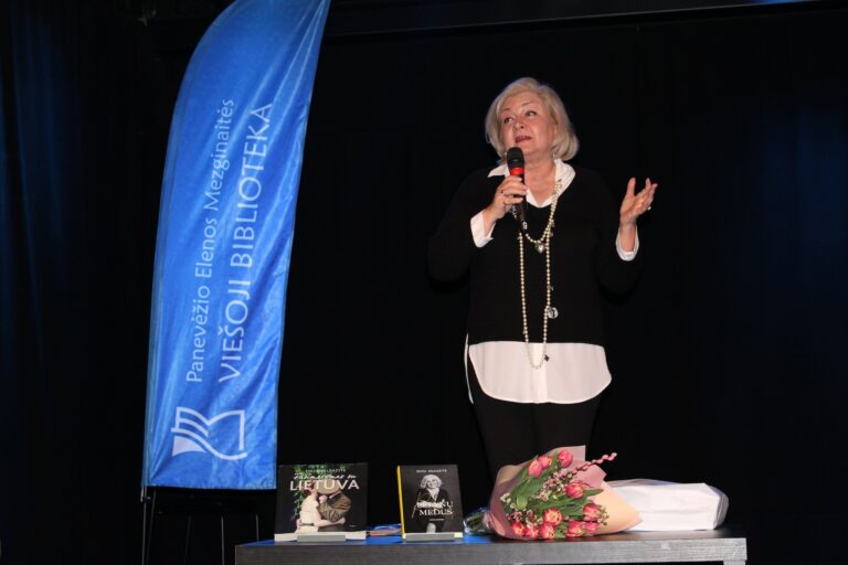 Scenoje stovi moteris su mikrofonu rankoje ir kalba auditorijai. Šalia jos – mėlyna vėliava su užrašu „Panevėžio Elenos Mezginaitės viešoji biblioteka“. Ant stalo priešais matomos dvi knygos ir gėlių puokštė. Tamsus fonas pabrėžia renginio atmosferą.