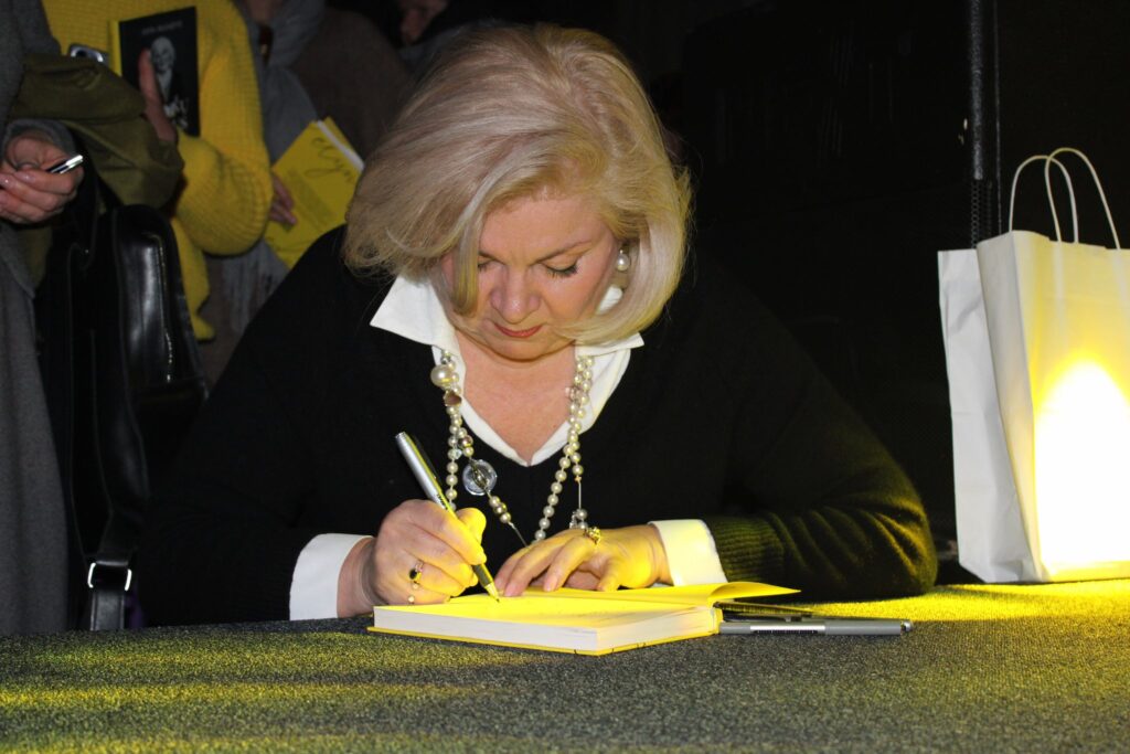 Edita Mildažytė sėdi prie stalo ir pasirašo knygoje, rankoje laiko rašiklį. Ant stalo padėta knyga, šalia matyti dar vienas rašiklis ir šviečiantis popierinis maišelis, fone stovi žmonės.