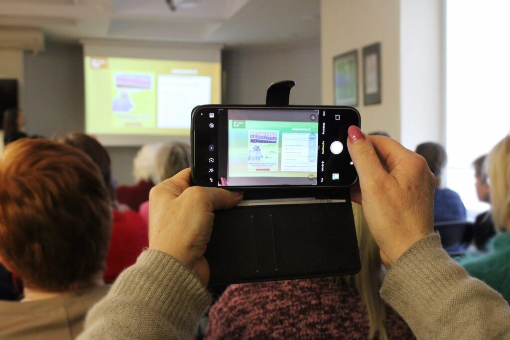 Asmuo rankose laiko išmanųjį telefoną ir fotografuoja pristatymo skaidrę projektoriaus ekrane, o priešais sėdi renginio ar paskaitos klausytojai.