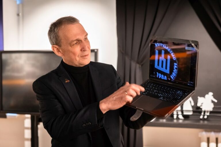 vyras laiko kompiuterį, vaizduojantį Gedimino stulpus, Miltinio kambario fone