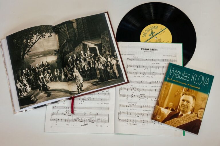 Atversta knyga, kurioje yra operos scena, išdėlioti atverstos natos, muzikos plokštelė, knygas su kompozitoriaus A. Klovos nuotrauka
