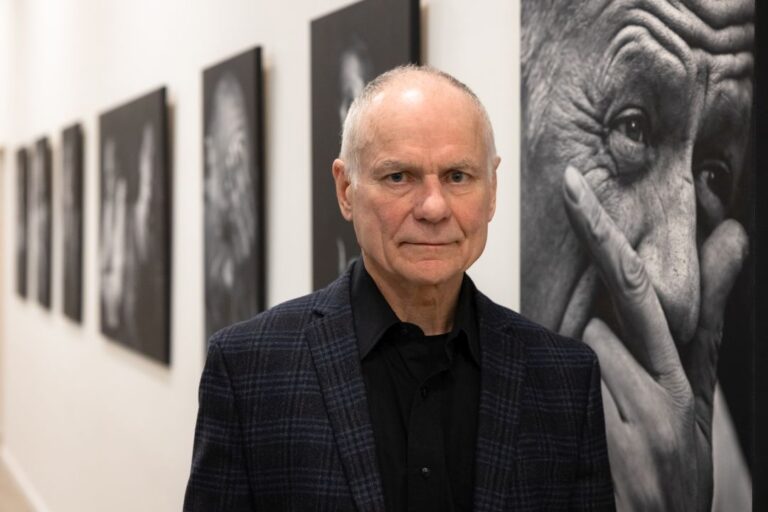 Vyras žvelgia rimtai stovėdamas juodai baltų nuotraukų ant baltų sienų fone