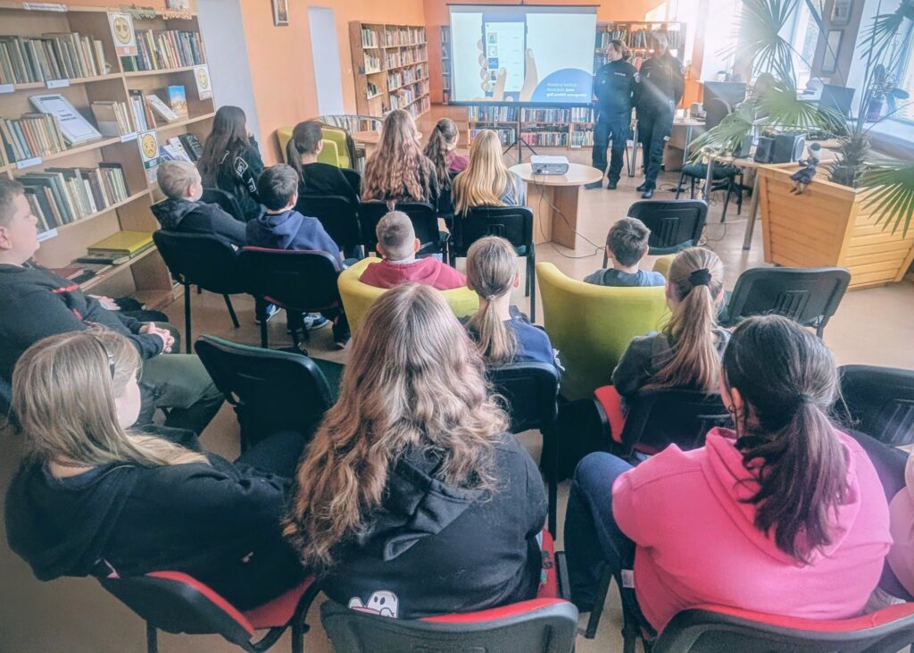 Paveikslėlyje matyti grupė jaunų žmonių, sėdinčių bibliotekoje ir stebinčių pristatymą. Priekyje rodomas skaidrės su ekranu, kuriame matoma telefone atvaizduota informacija. Pateikimą veda dvi policijos pareigūnės, kurios dalijasi informacija su jaunaisiais klausytojais.