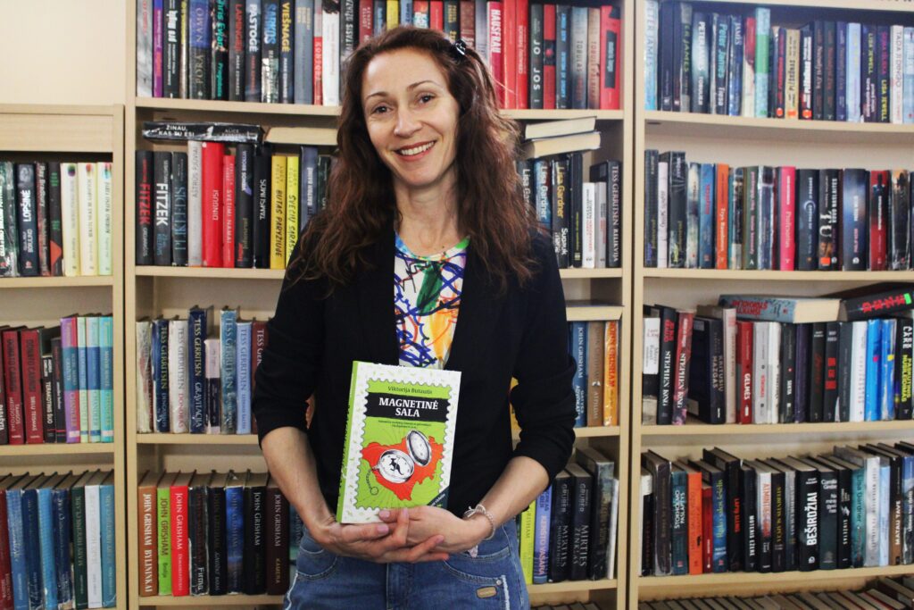 Moteris stovi priešais knygų lentyną ir šypsodamasi rankose laiko knygą „Magnetinė sala“, kurios viršelyje pavaizduotas kompasas ir Australijos kontūras.