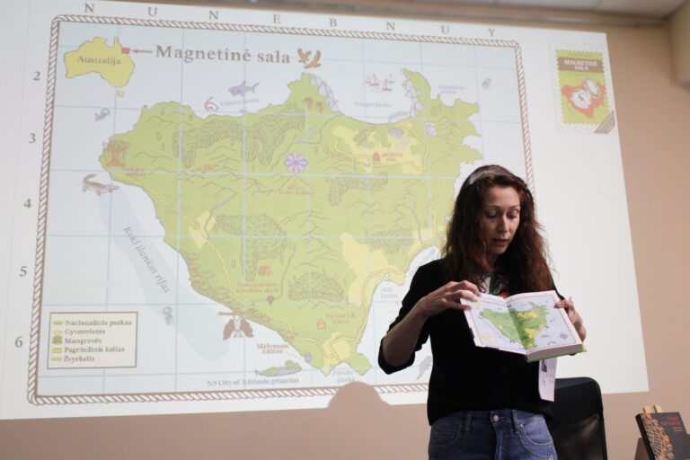 Moteris stovi priešais projekcijoje rodomą Australijos „Magnetinės salos“ žemėlapį ir rankose laiko atverstą knygą su tuo pačiu žemėlapiu, pristatydama medžiagą auditorijai.