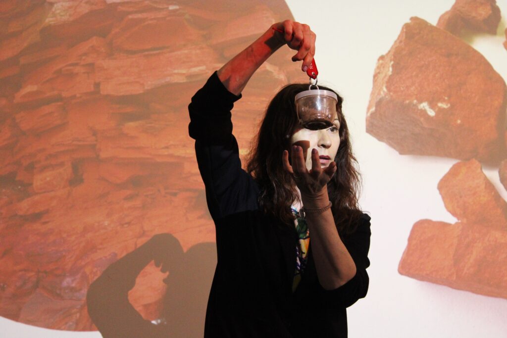 Moteris pristatymo metu pakėlusi ranką laiko mažą indelį su tamsiu turiniu, o už jos rodomoje projekcijoje matyti stambiai pavaizduotos uolienos.