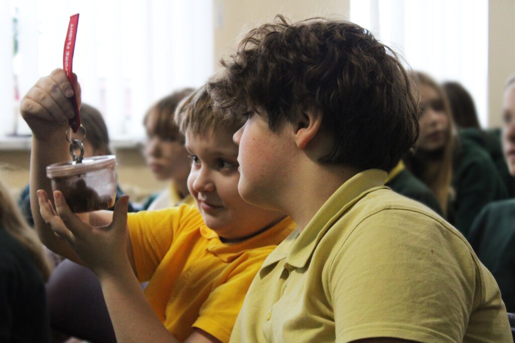 Matome du berniukus, sėdinčius klasėje ir žiūrinčius į stiklainį, kurį vienas iš jų laiko. Berniukas, laikantis stiklainį, rodo jį kitam, kuris su susidomėjimu žiūri. Abu berniukai dėvi geltonas marškinėlius, o fone matosi keli kiti vaikai.