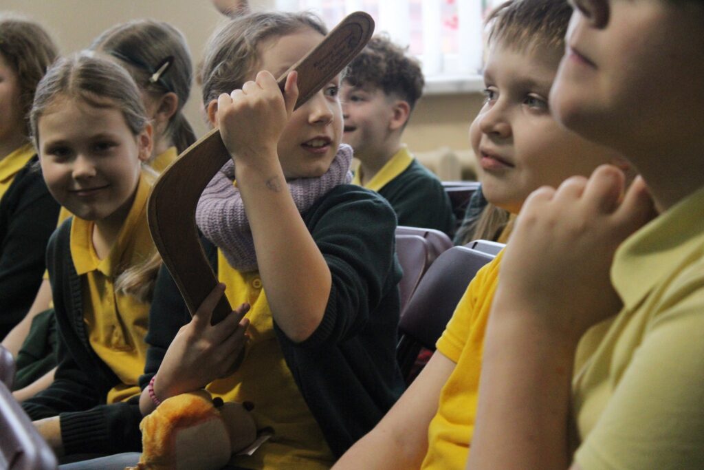 Matome grupę vaikų, sėdinčių auditorijoje. Viena mergaitė laiko boomerangą ir rodo jį šalia sėdinčiai draugei, kuri šypsosi. Vaikai dėvi geltonas marškinėlius ir žalius megztinius. Kai kurie vaikai žiūri į boomerangą su susidomėjimu, o kiti už jų šiek tiek matosi fone.