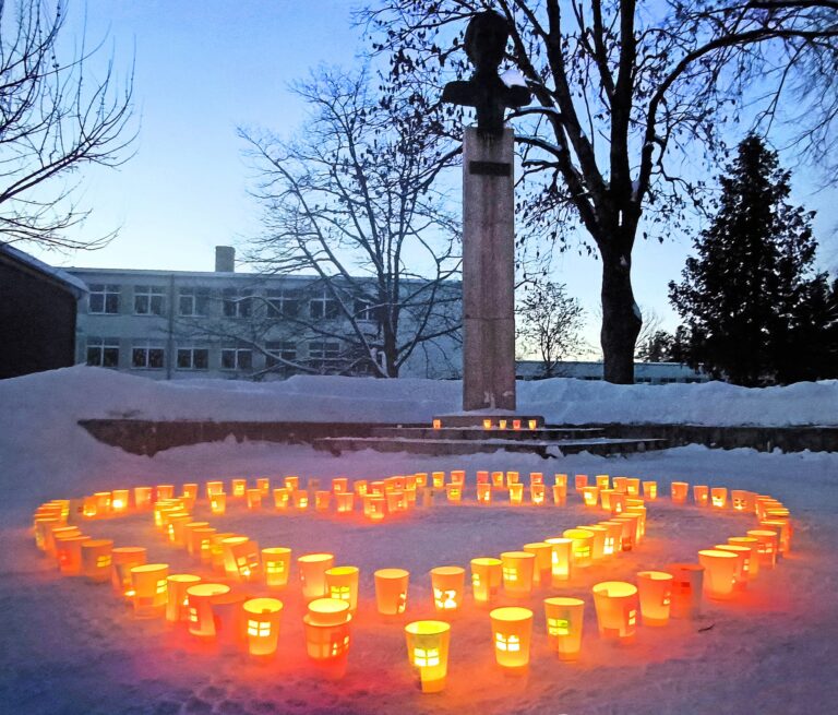 Žiemos vakare sniege sustatytos degančios žvakės sudaro širdies formą. Fone matyti paminklas su biustu ir medžiai be lapų, aplink sniego pusnys bei pastatas.
