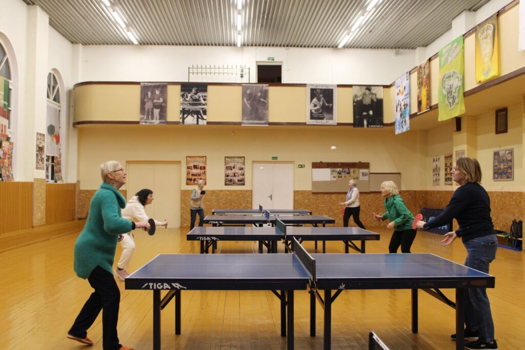 Sporto salėje keli vyresnio amžiaus žmonės žaidžia stalo tenisą prie trijų stalų. Priekyje dvi moterys apsikeičia smūgiais, kiti dalyviai žaidžia fone. Ant sienų kabo plakatai, grindys medinės, apšvietimas ryškus.