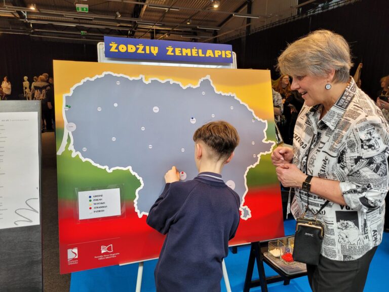 Vaikas ir vyresnio amžiaus moteris stovi prie interaktyvaus stendo su Lietuvos žemėlapiu „Žodžių žemėlapis“. Vaikas prisega žymeklį prie žemėlapio, o moteris stebi ir šypsosi. Stendas spalvotas Lietuvos vėliavos spalvomis, šalia matyti legenda su skirtingų žymėjimų paaiškinimais.