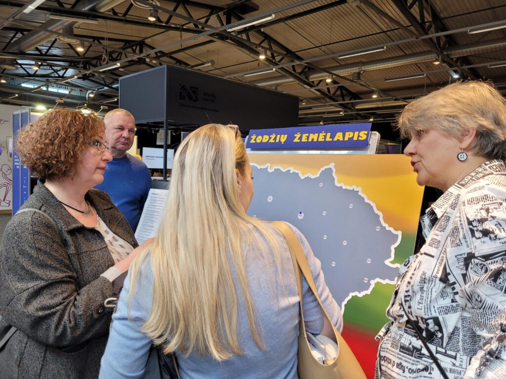 Keturi suaugusieji stovi prie stendo su užrašu „Žodžių žemėlapis“ ir Lietuvos žemėlapiu. Trys žmonės žiūri į žemėlapį iš arti, o moteris dešinėje, vilkinti raštuotus marškinius, kalba ir rodo į stendą. Stendas stovi parodų salėje su aukštomis lubomis ir kitais stendais fone.