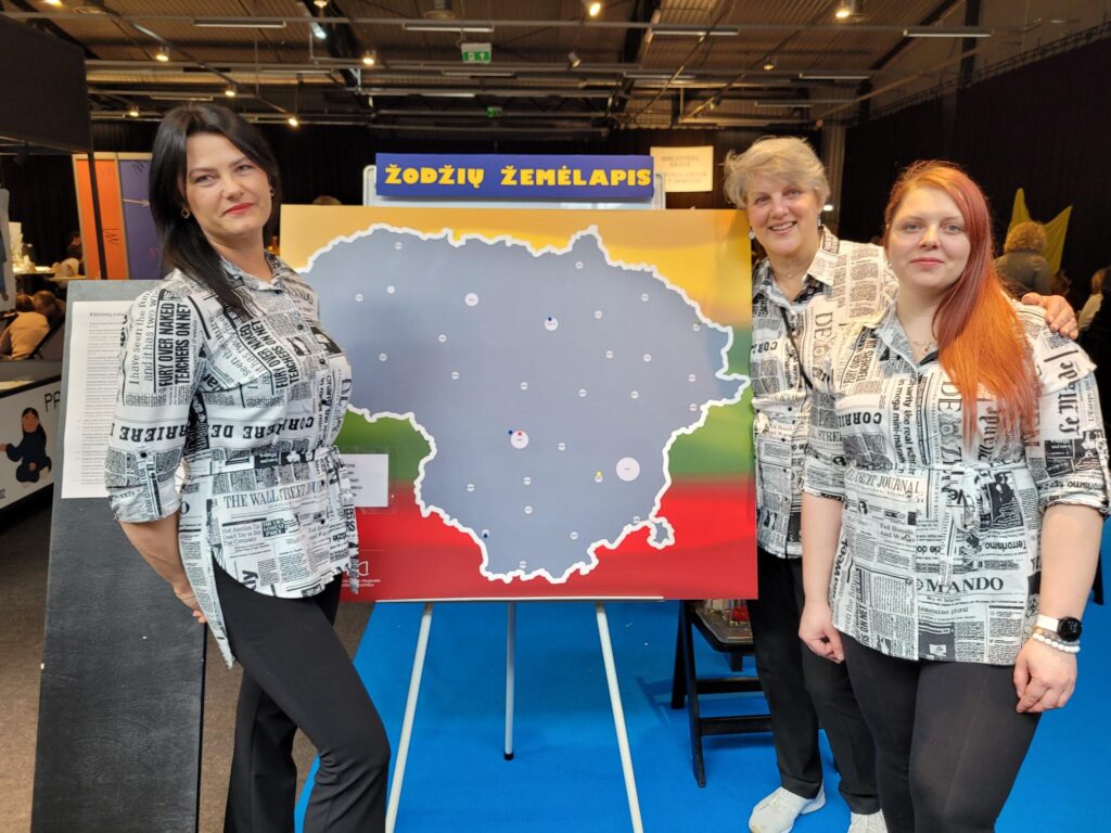Trys moterys stovi parodų salėje prie stendo su užrašu „Žodžių žemėlapis“ ir Lietuvos žemėlapiu. Jos vilki vienodus, laikraščių raštais margintus marškinius ir šypsodamosi pozuoja nuotraukai. Fone matyti renginio erdvė su kitais stendais ir aukštomis lubomis.