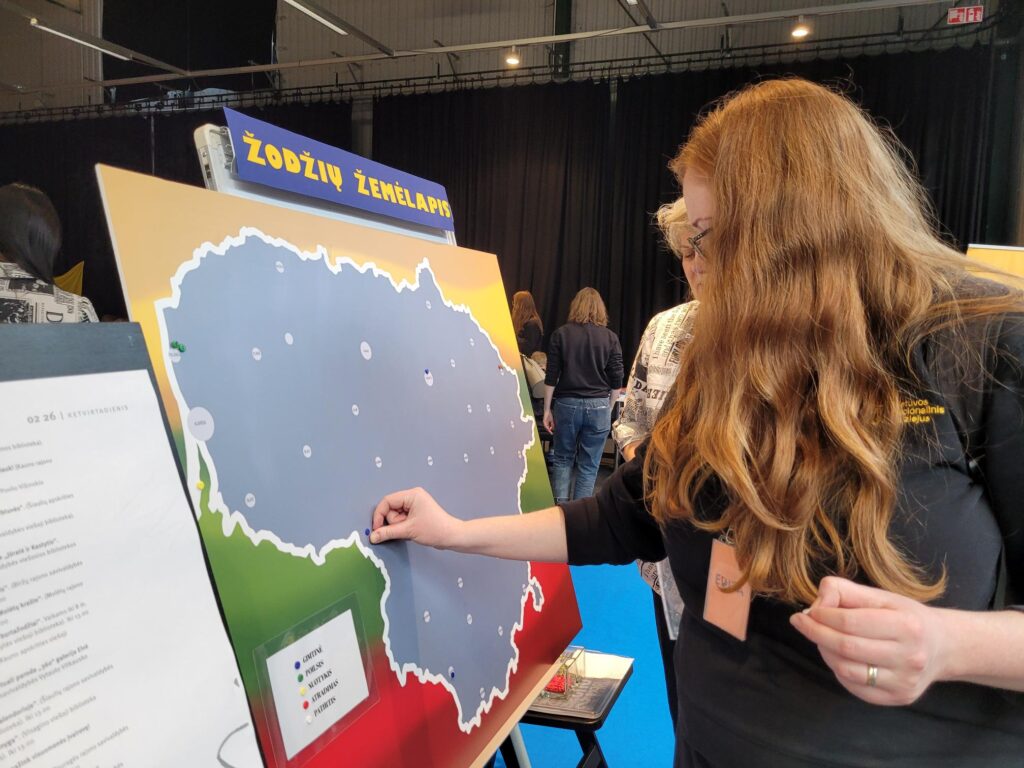 Moteris stovi prie stendo su Lietuvos žemėlapiu ir smeigia smeigtuką į pasirinktą vietą. Viršuje matyti užrašas „Žodžių žemėlapis“, aplink – parodos erdvė su kitais žmonėmis.