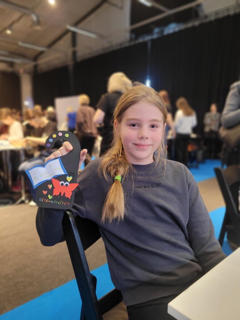 Mergaitė sėdi renginio erdvėje ir šypsodamasi laiko rankoje savo pagamintą juodą durų pakabuką su spalvingomis detalėmis, užrašu „Netrukdykite!!!“ ir piešiniu. Fone matyti kiti žmonės ir vykstančios veiklos stalai.