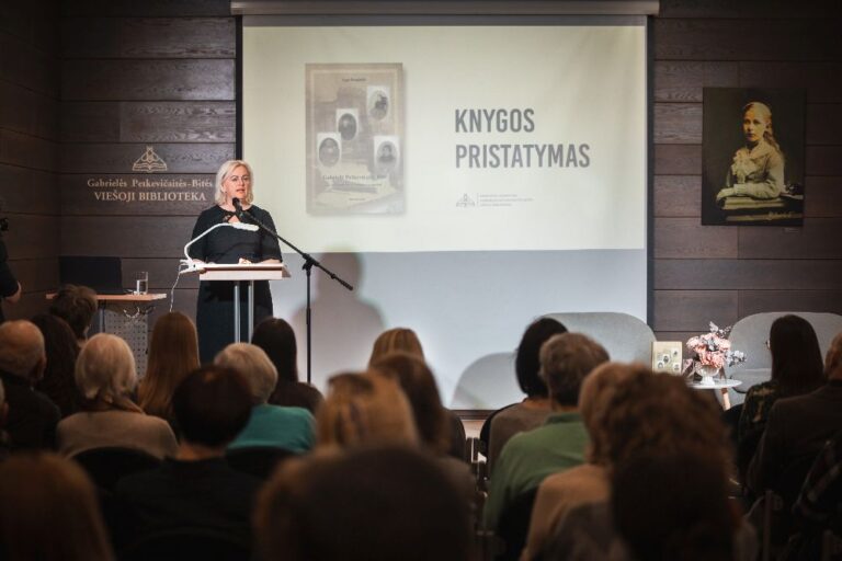 Moteris knygos autorė stovi priešais auditoriją prie mikrofono ant pakylos bibliotekos salės fone