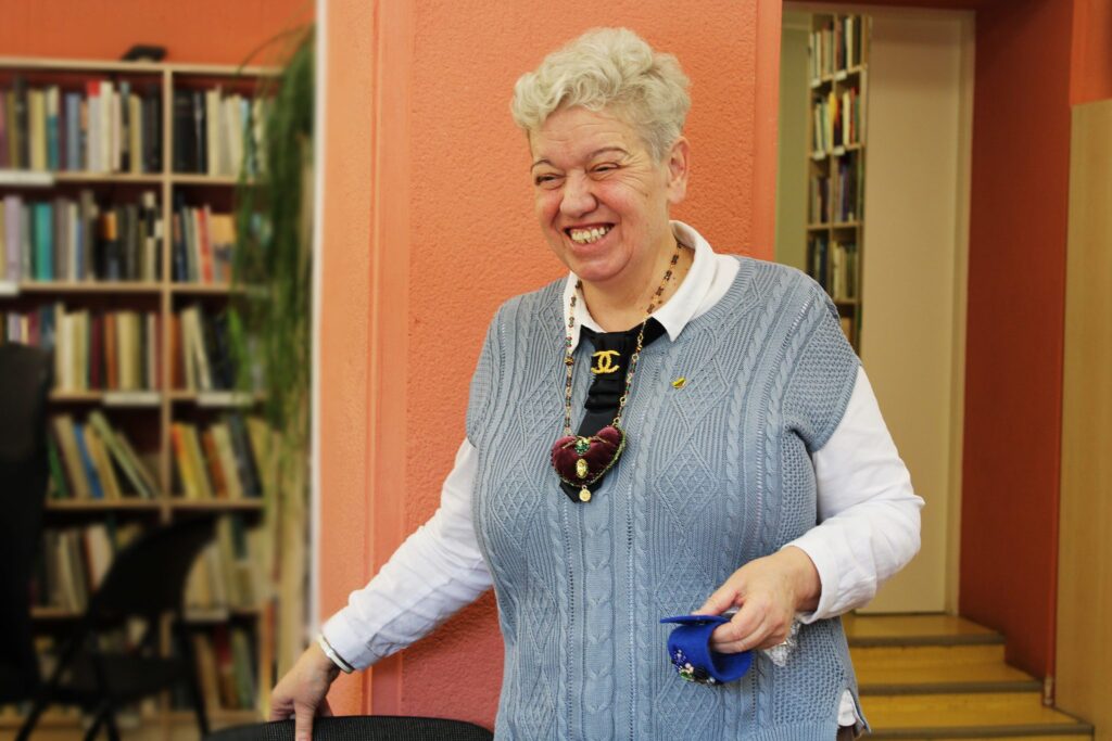 Bibliotekos erdvėje stovi besijuokianti moteris su pilka megzta liemene ir baltais marškiniais, seginti didelį rankų darbo kaklo papuošalą su širdelės formos pakabuku.