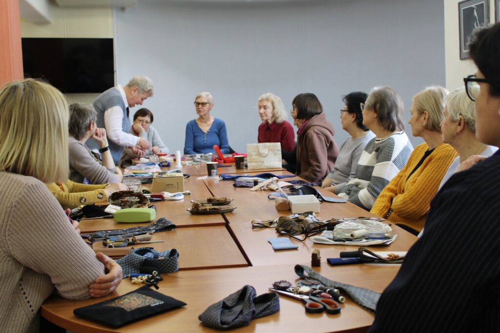 Kūrybinių dirbtuvių metu žmonių grupė sėdi prie stalo, klausosi vedėjos ir kuria rankų darbo kaklo papuošalus iš įvairių medžiagų.