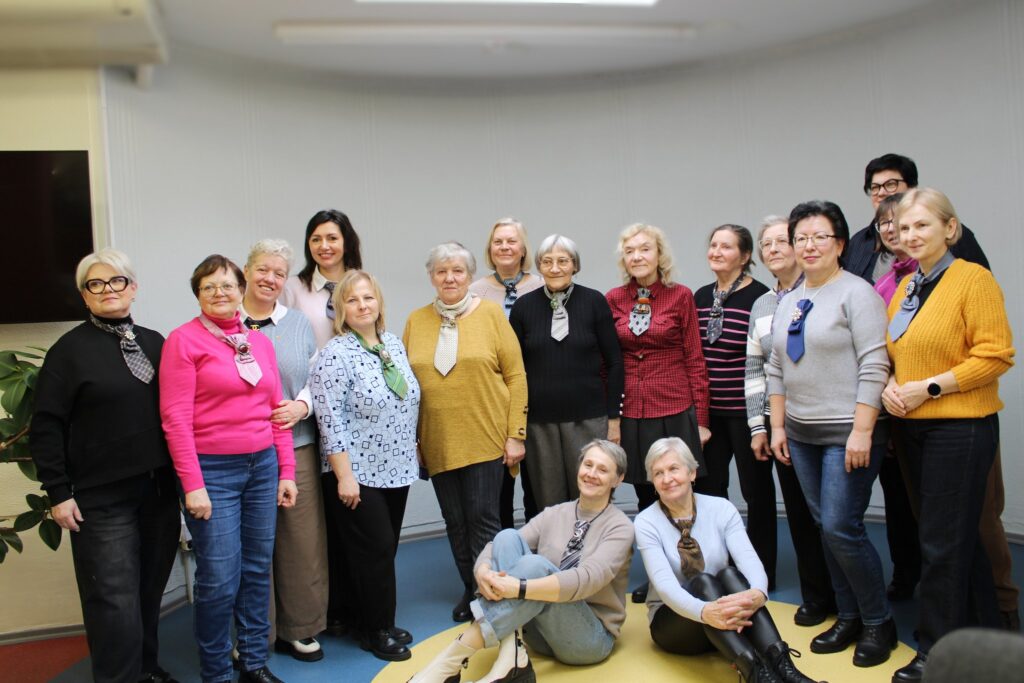 Grupė moterų stovi kartu ir pozuoja bendrai nuotraukai po kūrybinių dirbtuvių, daugelis jų segi iš kaklaraiščių pagamintus rankų darbo papuošalus.