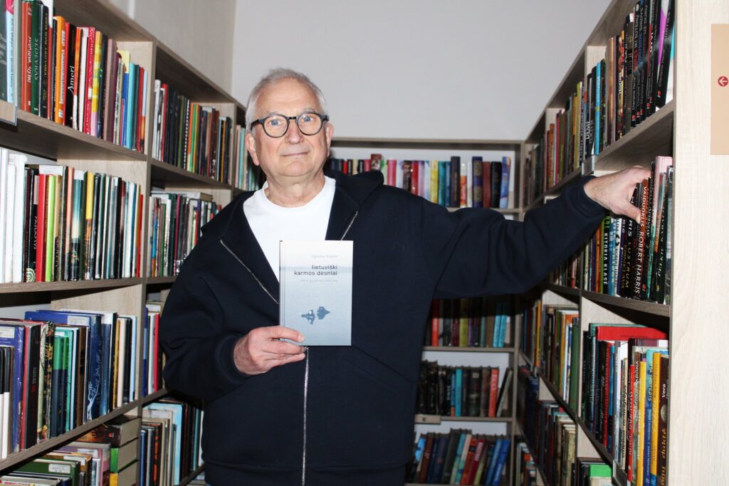 Nuotraukoje matomas vyras, laikantis knygą "Lietuviški karmos dėsniai" ir stovintis bibliotekos tarp knygų lentynų. Jis vilki tamsų džemperį su užtrauktuku ir šypsosi, rodydamas knygą. Už jo matoma daug kitų knygų ant lentynų.