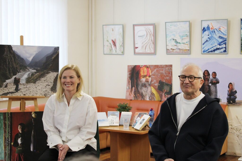 Nuotraukoje matomi vyras ir moteris, sėdintys ant kėdžių. Moteris šypsosi ir dėvi baltus marškinius, o vyras, apsirengęs tamsiu džemperiu, taip pat šypsosi. Už jų matomos įvairios nuotraukos.