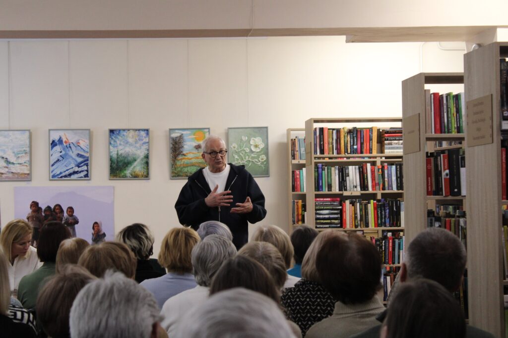 Nuotraukoje matomas vyras, kalbantis prieš auditoriją, stovintis bibliotekoje. Už jo yra nuotraukos ir meno darbai, o jis pats gestikuliuoja rankomis, aiškindamas temą. Pirmame plane matoma klausytojų grupė. Jų žvilgsniai nukreipti į kalbėtoją.