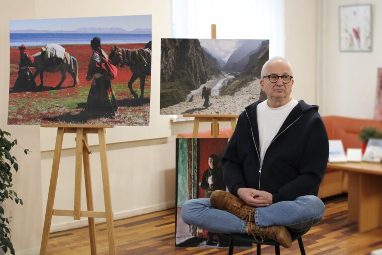 Nuotraukoje matomas vyras, sėdintis ant kėdės, sukryžiavęs kojas ir atsirėmęs abiem pėdomis į kėdės sėdynę. Jis turi atsipalaidavusią pozą, rankas padėjęs ant kelių. Jo apranga sudaro mėlynas džemperis su užtrauktuku, balta marškinėliais ir džinsai. Už jo matomos nuotraukos, vaizduojančios žmones, einančius su arkliais per raudoną žemę ir kalnų slėnį su upe.