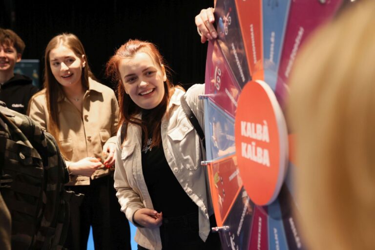 Trys besišypsantys jauni žmonės stovi prie žaidimo rato, viena mergina jį suka