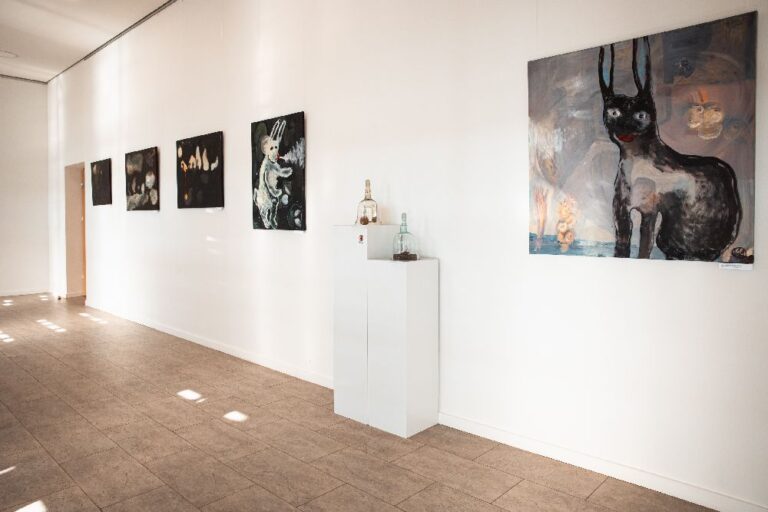 Ant baltos sienos kabantys kvadratiniai tapybos darbai ir du ant stovų stovintys stiklo ir sidabro parodos eksponatai bibliotekos koridoriaus fone