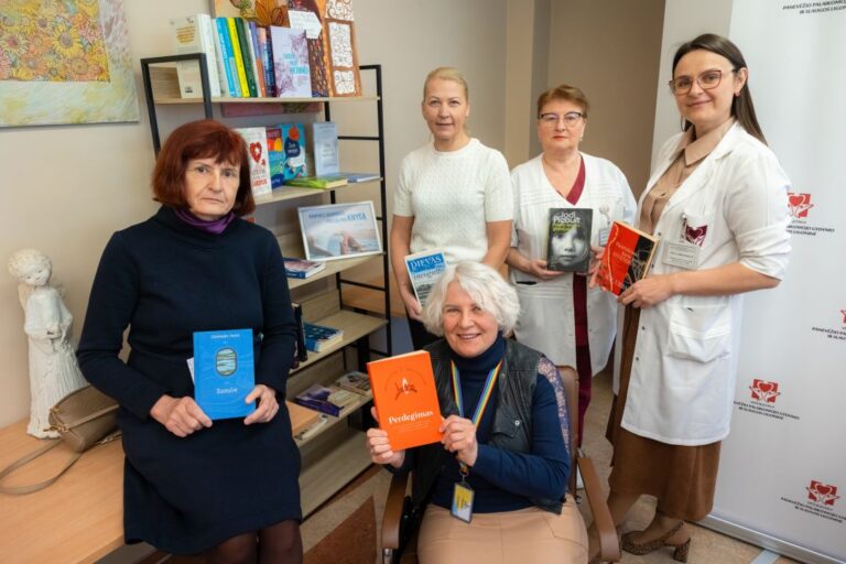 Šypsosi penkios moterys su knygomis rankose knygų lentynos ir patalpos fone