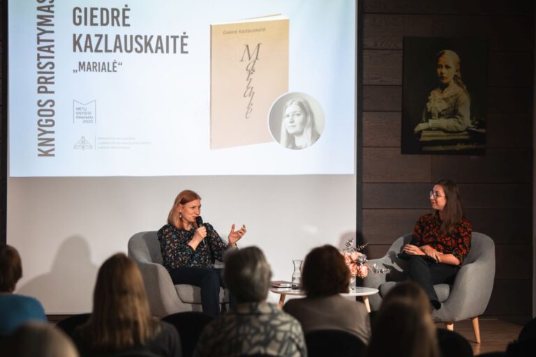 Dvi moterys sėdi ant pakylos priešais auditoriją salės fone