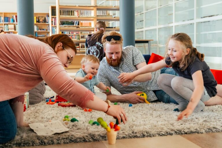 Dviejų suaugusiųjų ir dviejų vaikų šeima ant kilimo žaidžia ir dėlioja kaladėles bibliotekos salės fone