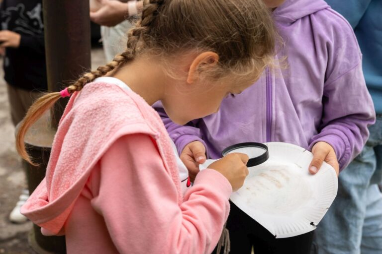 Mergaitė per didinamąjį stiklą tyrinėjanti baltą daiktą su užrašu kitų vaikų apsuptyje