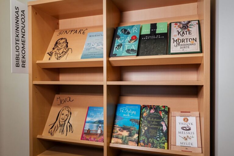 Knygų lentynose išdėliotos knygos ir užrašas Bibliotekininkas rekomenduoja