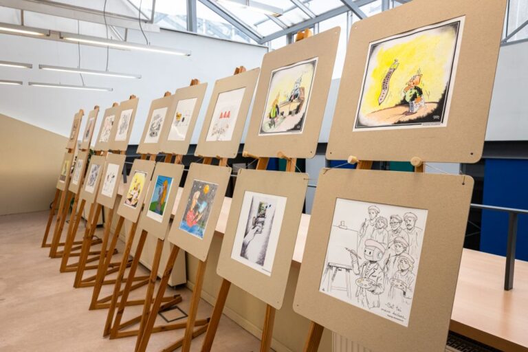 Ant stovų išdėliota karikatūrų paroda bibliotekos erdvės fone