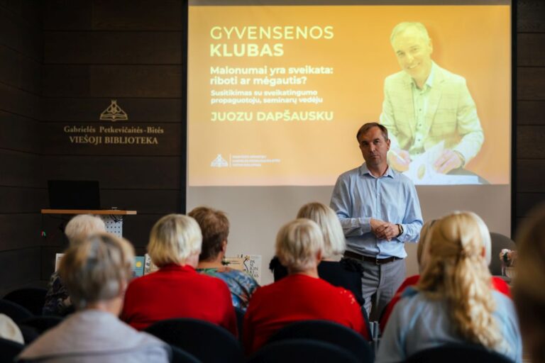 Gyvensenos klubo lektorius stovi priešais renginio auditoriją projektoriaus ir salės fone