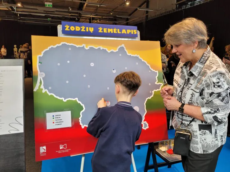 Vaikas ir vyresnio amžiaus moteris stovi prie interaktyvaus stendo su Lietuvos žemėlapiu „Žodžių žemėlapis“. Vaikas prisega žymeklį prie žemėlapio, o moteris stebi ir šypsosi. Stendas spalvotas Lietuvos vėliavos spalvomis, šalia matyti legenda su skirtingų žymėjimų paaiškinimais.
