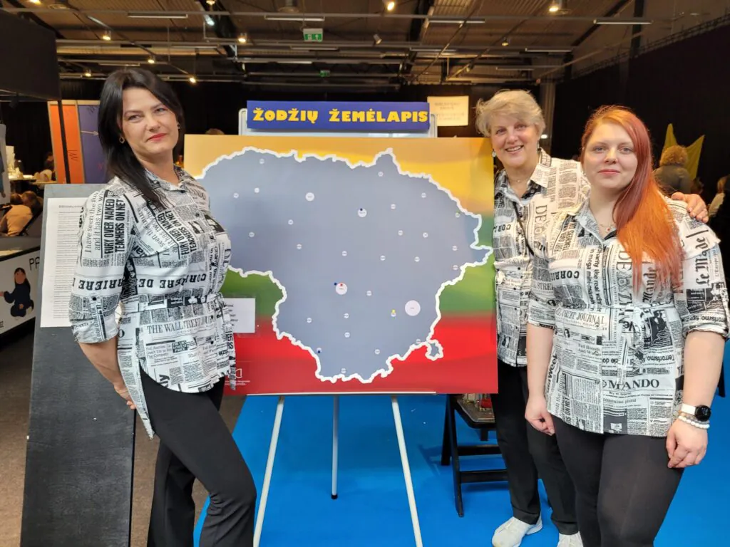 Trys moterys stovi parodų salėje prie stendo su užrašu „Žodžių žemėlapis“ ir Lietuvos žemėlapiu. Jos vilki vienodus, laikraščių raštais margintus marškinius ir šypsodamosi pozuoja nuotraukai. Fone matyti renginio erdvė su kitais stendais ir aukštomis lubomis.