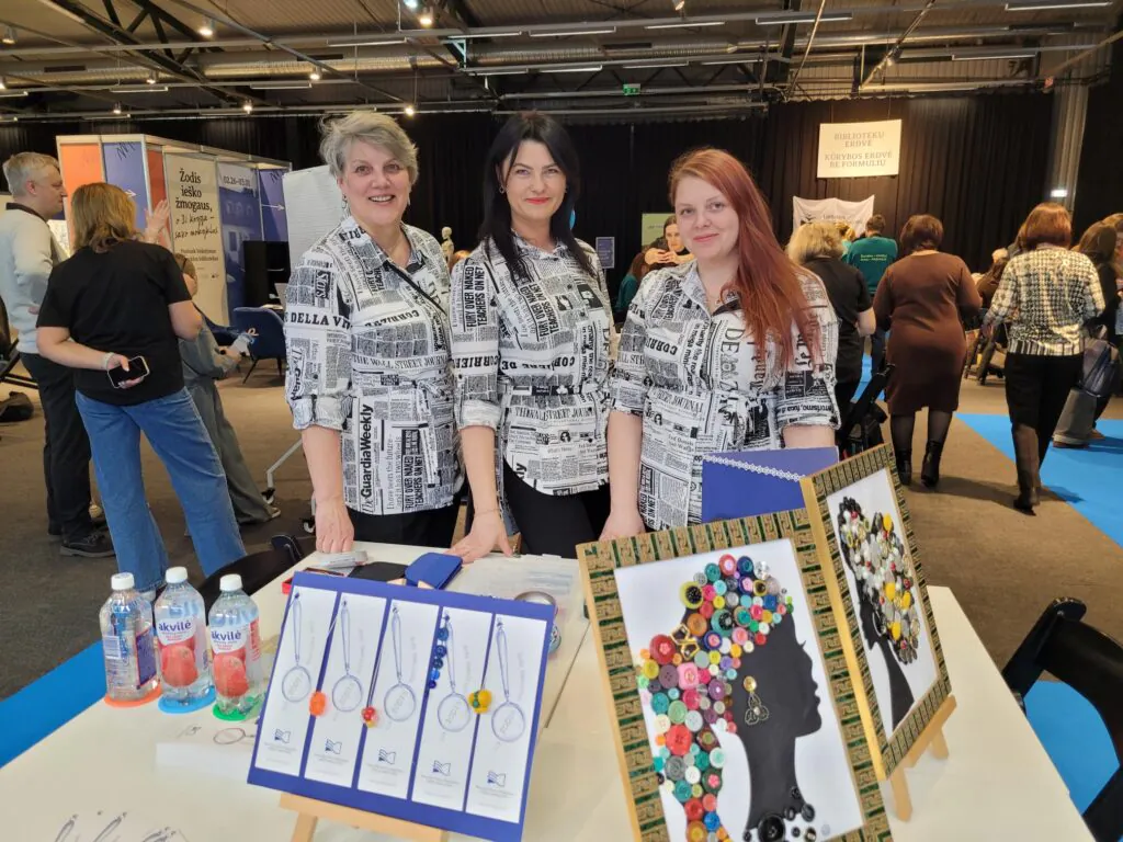 Trys moterys stovi už stendo renginio erdvėje ir šypsosi į kamerą. Ant stalo priešais jas – kūrybiniai darbai: rėmeliuose įrėminti paveikslai su iš sagų sudėliotais moters siluetais, knygų skirtukai ir vandens buteliukai. Aplink matyti kiti renginio dalyviai ir informaciniai stendai.